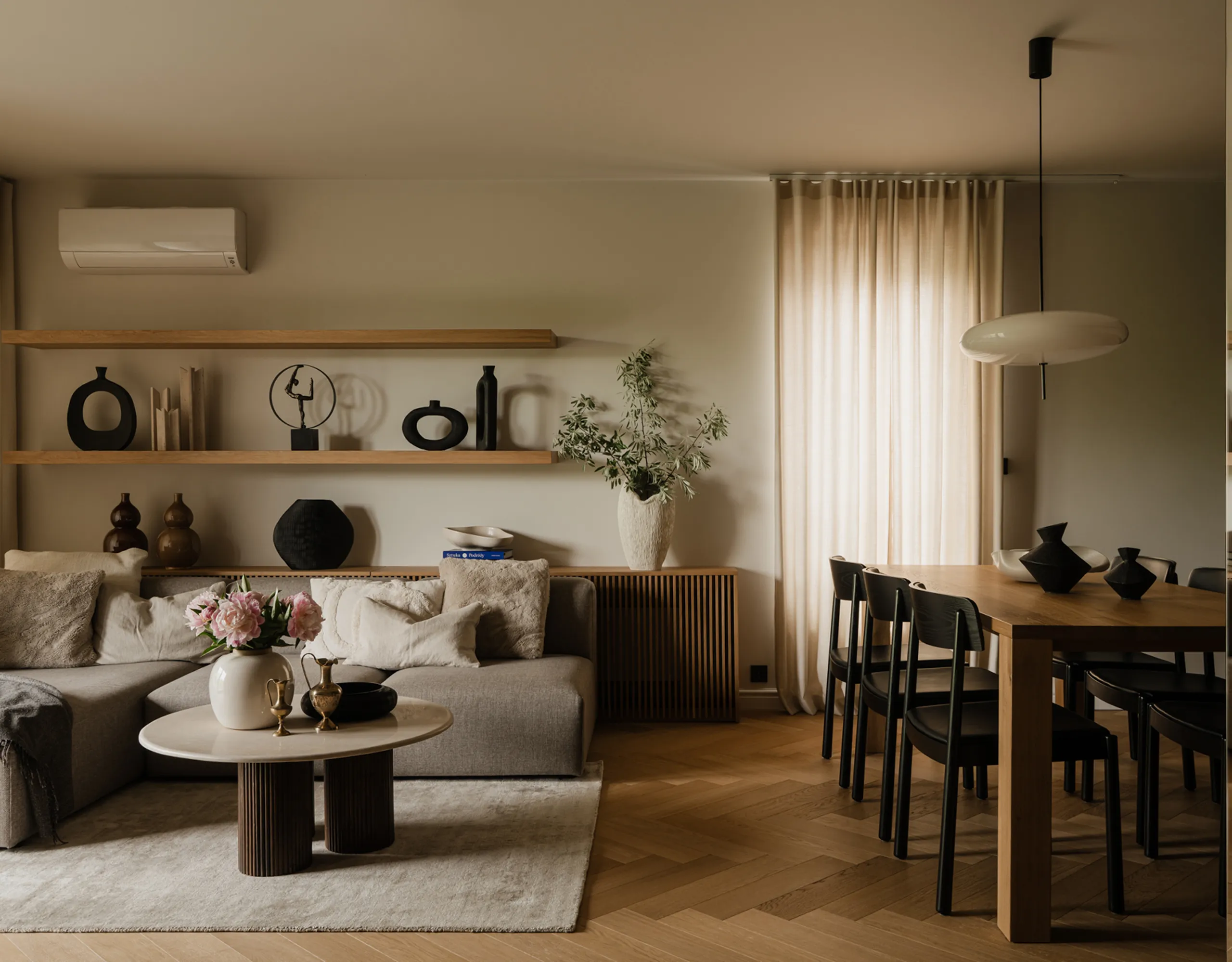 Elegancki salon z beżową sofą narożną, okrągłym stolikiem i drewnianym stołem jadalnym - NAM STUDIO